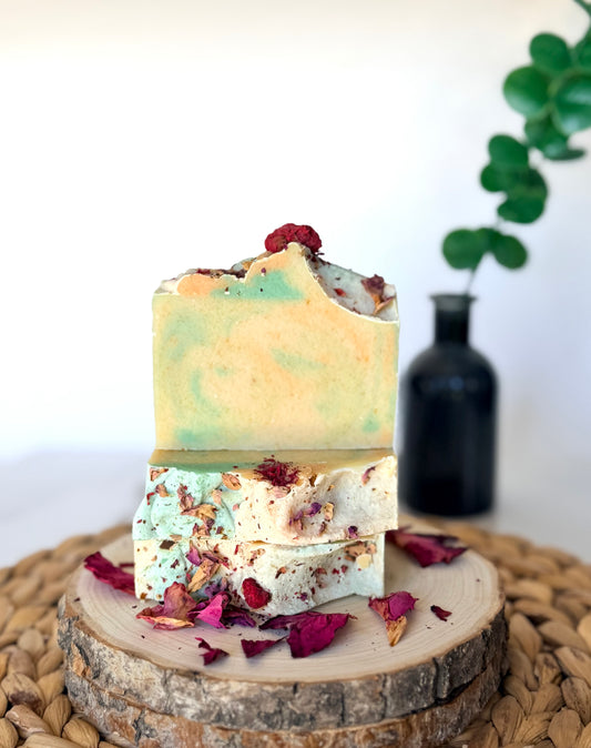 Two handmade soap bars with soft green and golden swirls, topped with dried botanicals and rose petals on a natural wood slice.