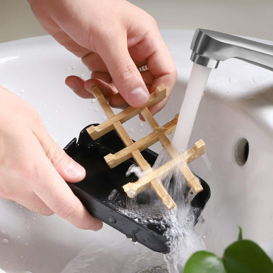 Black draining soap dish being rinsed under running water with the removable bamboo rack lifted out for easy cleaning.