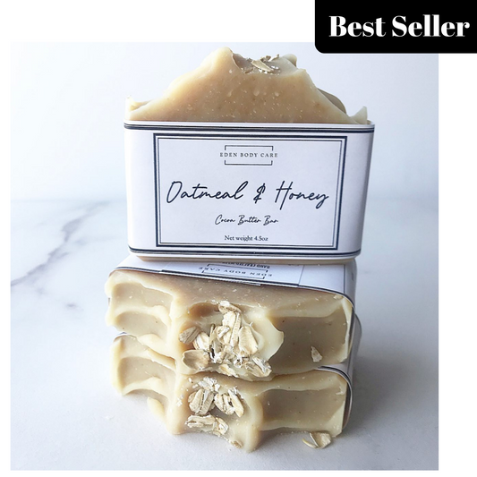 Stack of beige oatmeal and honey goat milk soap bars with oatmeal pieces on top, placed on a white surface with a paper cigar band packaging.