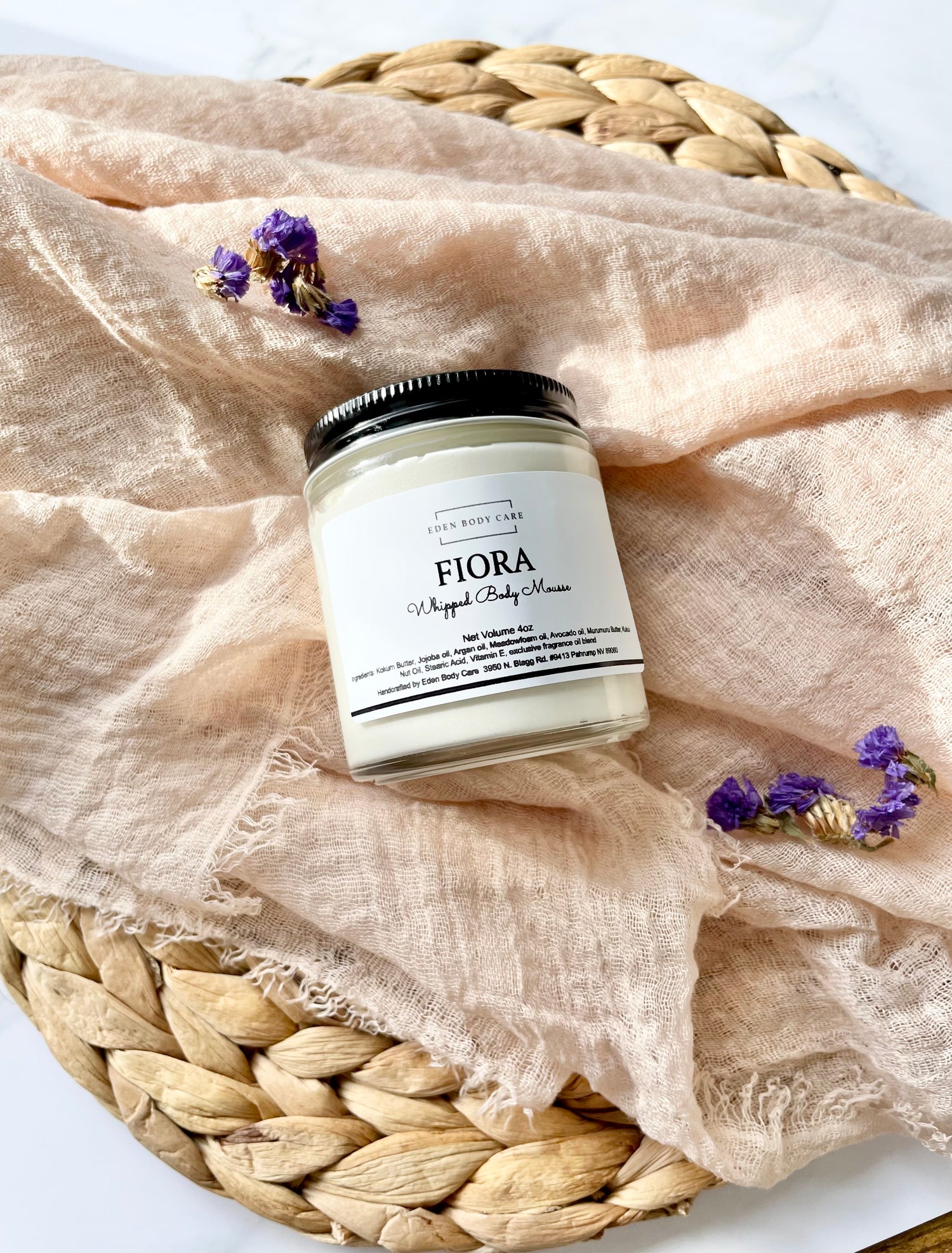 Eden Body Care Fiora whipped body mousse in a glass jar displayed on soft neutral fabric with small purple flowers.