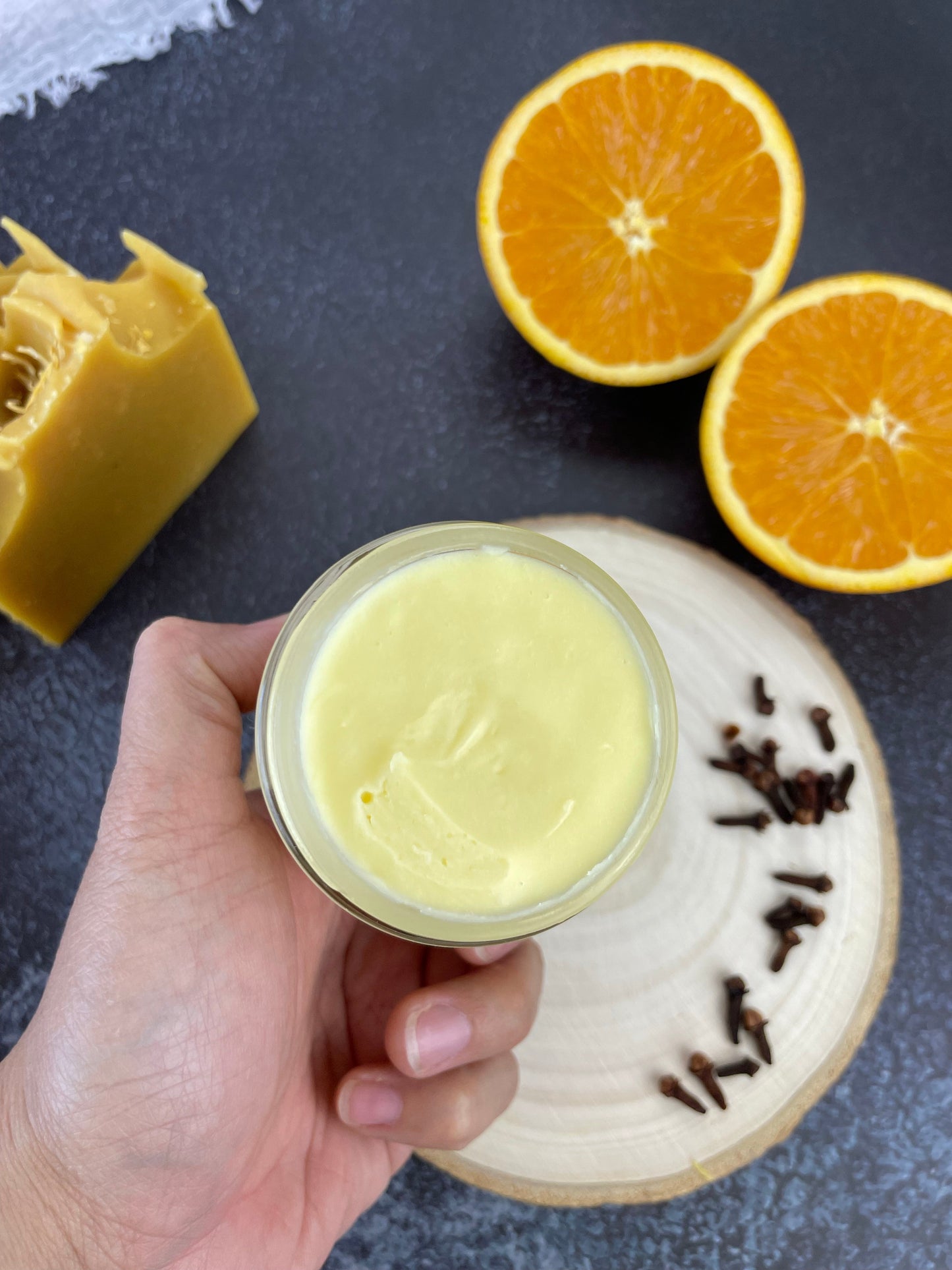 Open jar of Eden Body Care Orange & Clove body butter held in hand, showing its smooth whipped texture beside fresh orange halves, whole cloves, and a matching soap bar.