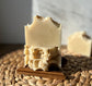 A bar of Oatmeal Vegan Hemp Milk Soap is partially sliced, revealing its creamy white interior with visible flakes of oats. The soap is placed on a woven mat, with a wooden piece underneath to support it.