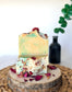 Two handmade soap bars with soft green and golden swirls, topped with dried botanicals and rose petals on a natural wood slice.