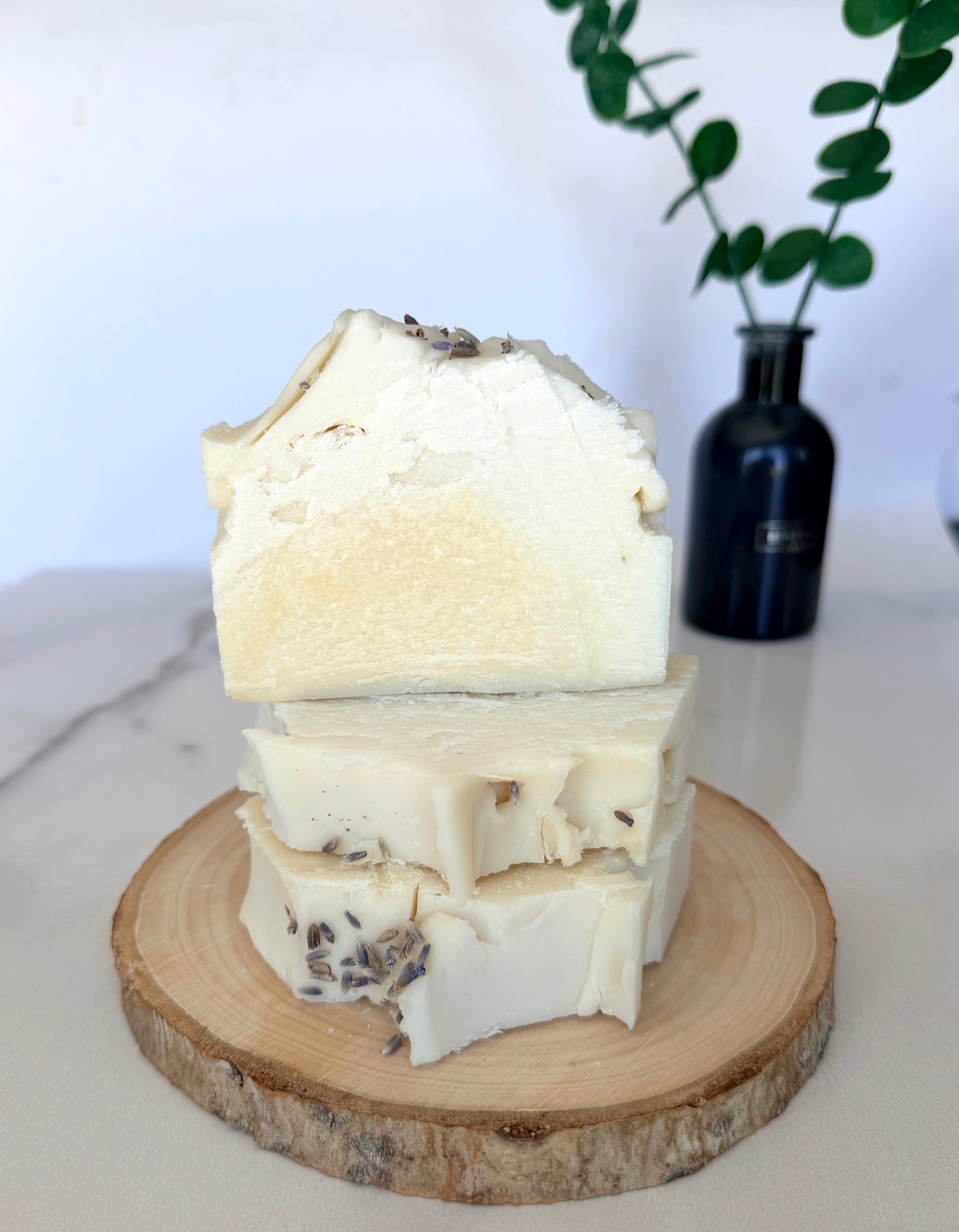 Stack of handmade lavender lemon goat milk soap bars displayed on a natural wood slice with a black vase and greenery in the background.