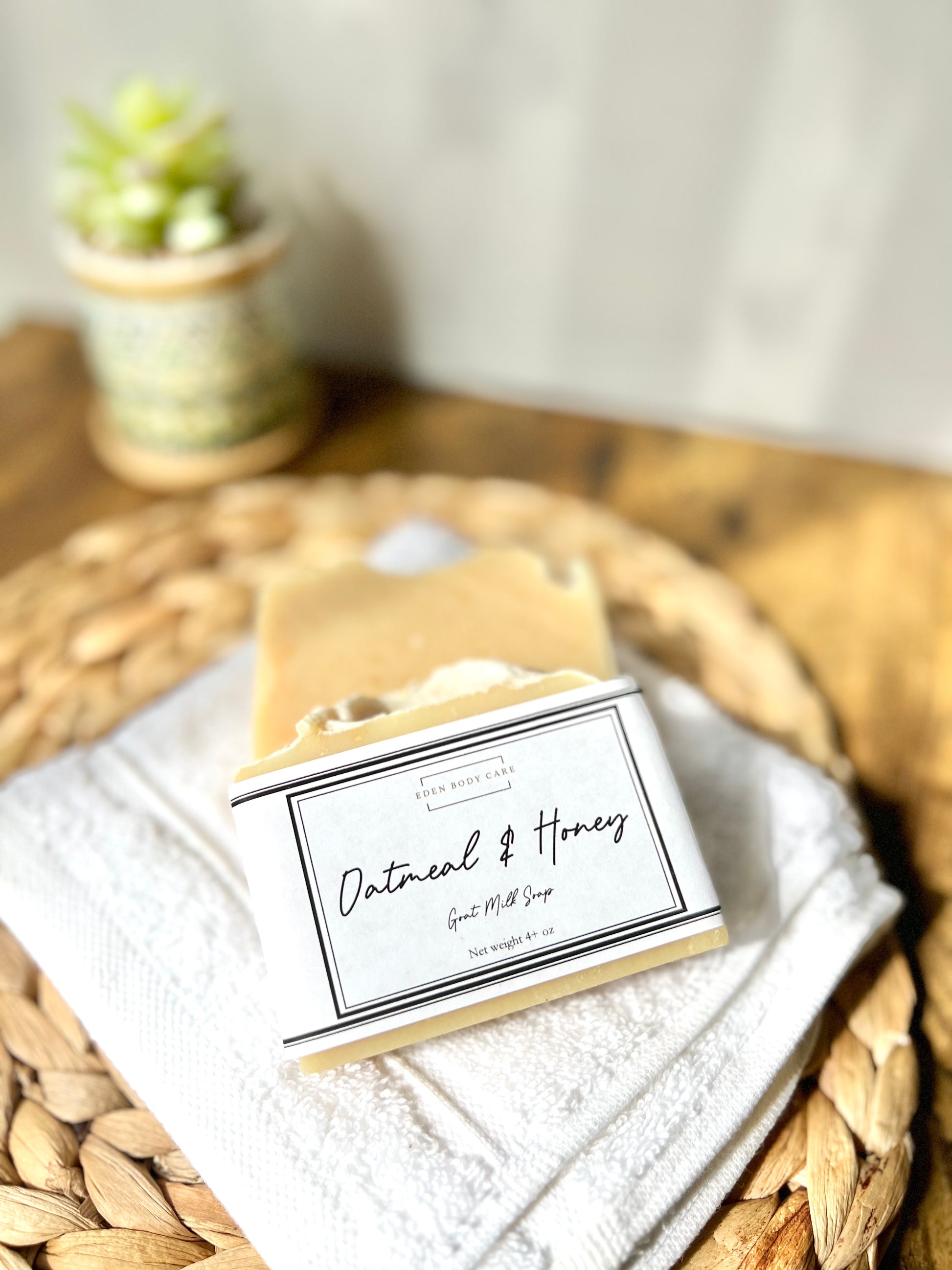 Stack of beige oatmeal and honey goat milk soap bars with oatmeal pieces on top, placed on a white wash cloth with a paper cigar band packaging.