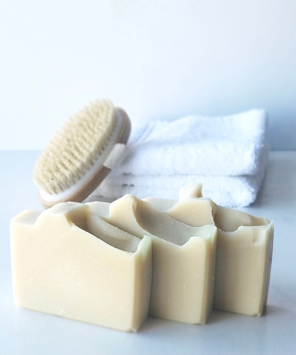 Three Eden Body Care Classic Unscented goat milk soap bars displayed beside a natural bath brush and folded white towels
