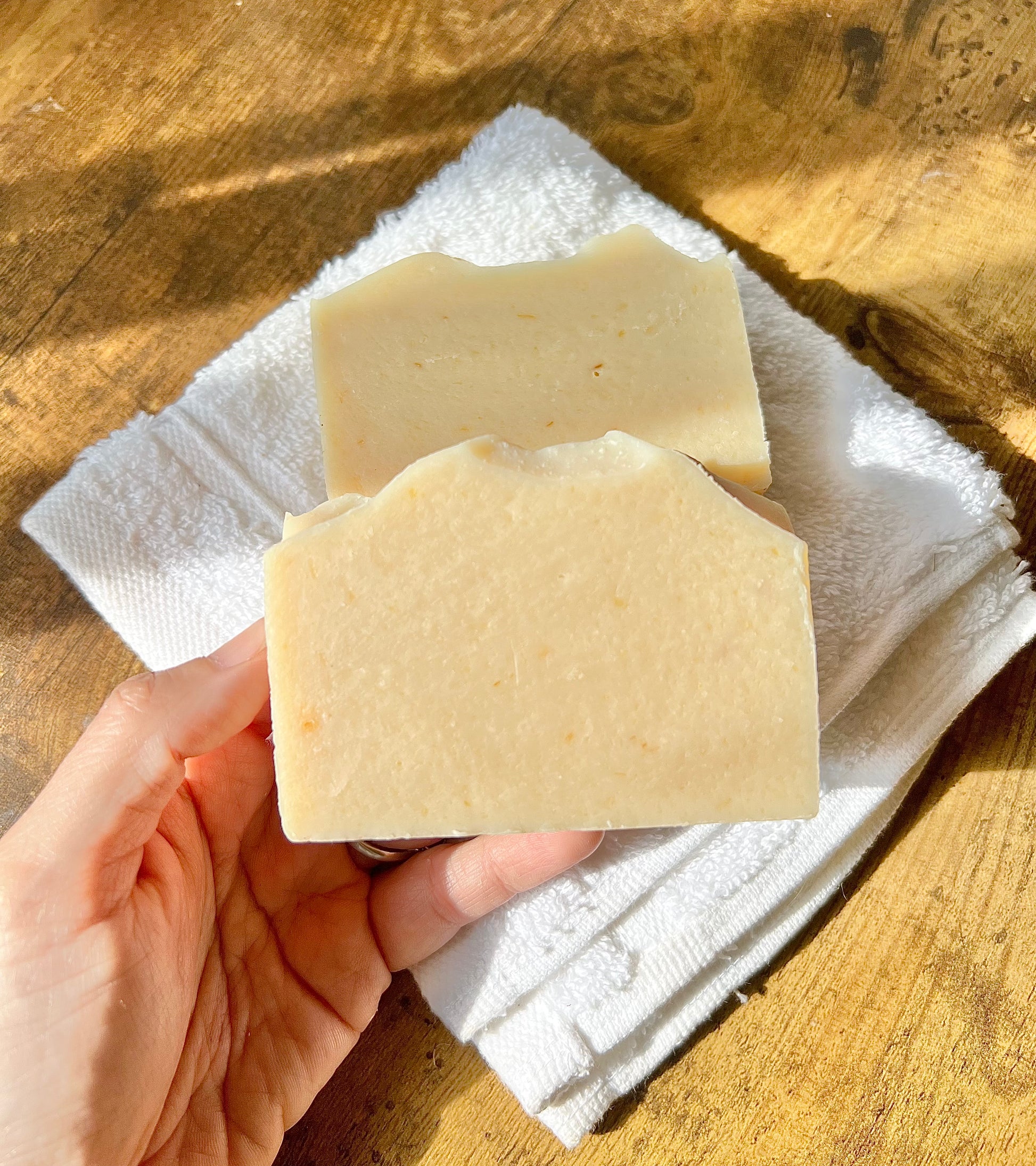 Eden Body Care Classic Unscented goat milk soap bar held in hand over a folded white towel in warm natural sunlight