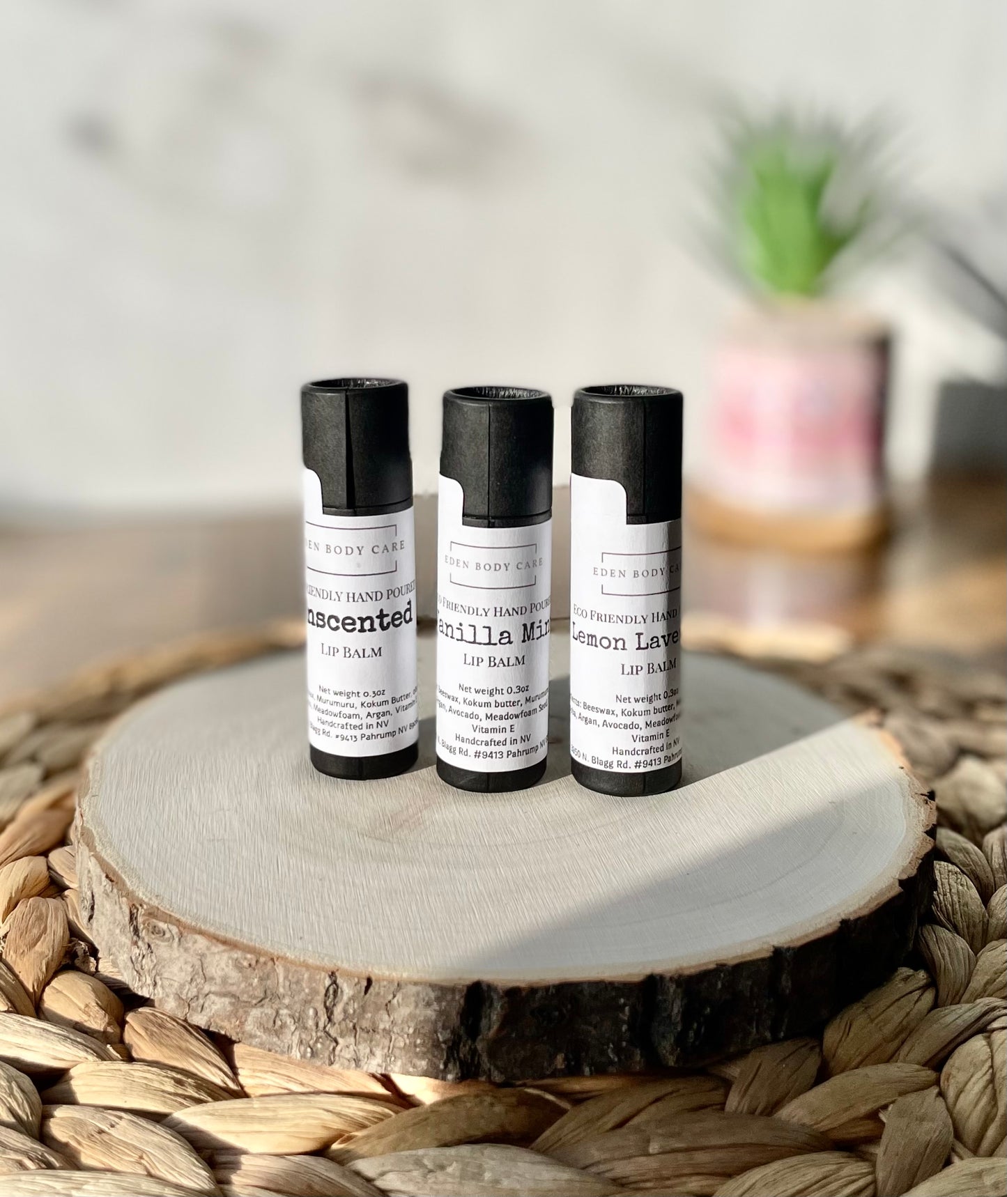 Three Eden Body Care lip balm tubes in Unscented, Vanilla Mint, and Lemon Lavender displayed together on a natural wood slice.