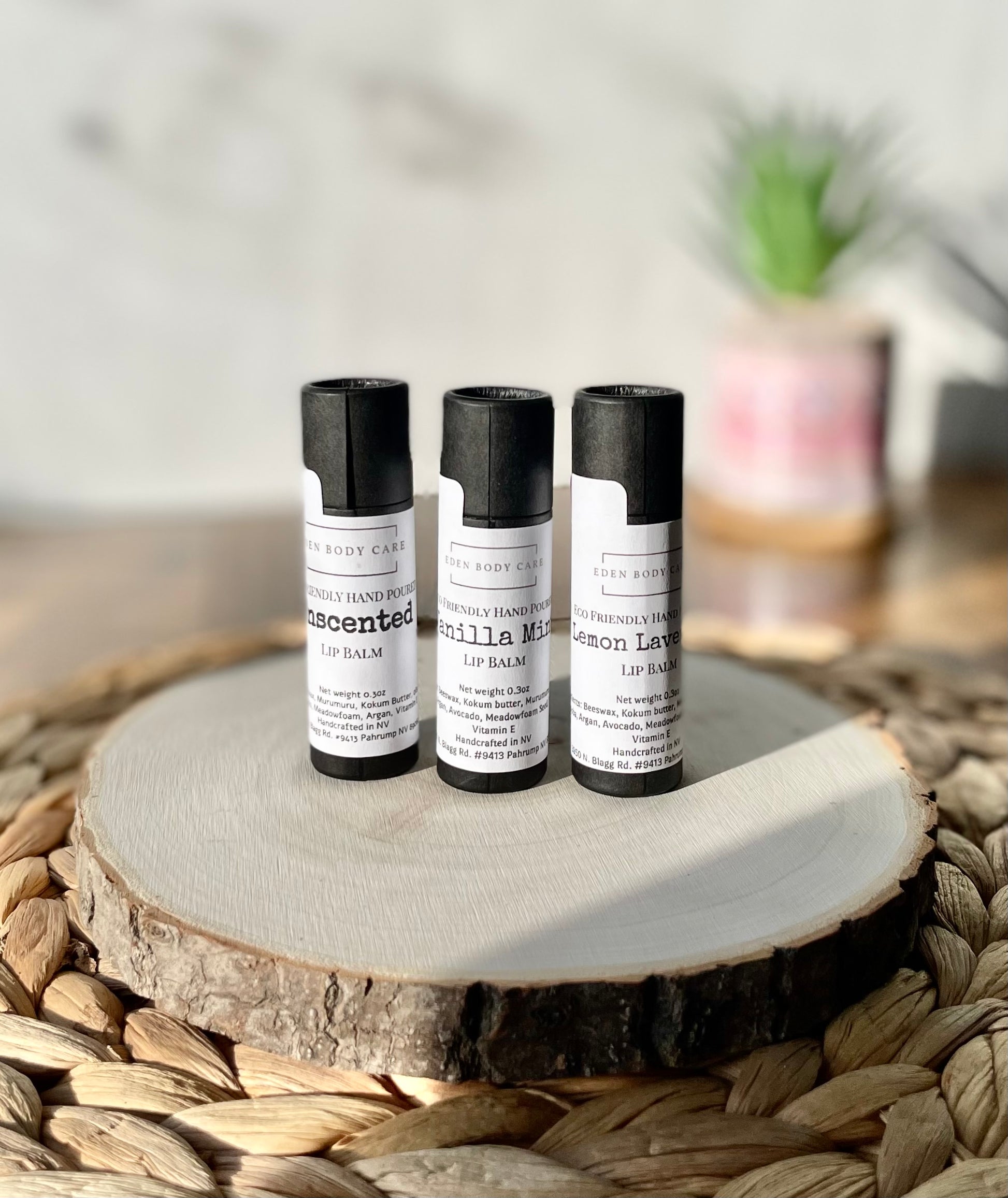 Three Eden Body Care lip balm tubes in Unscented, Vanilla Mint, and Lemon Lavender displayed together on a natural wood slice.