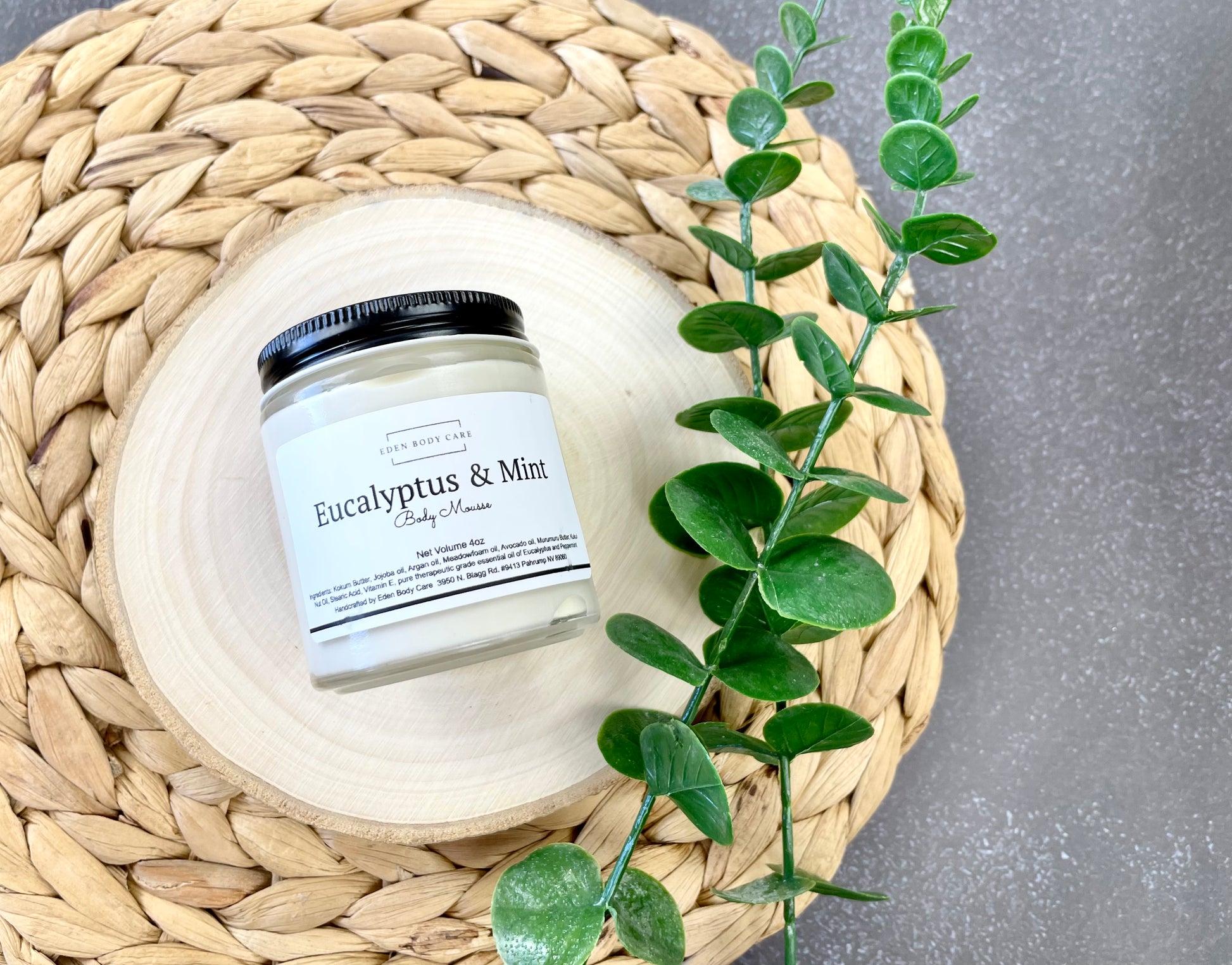 Eden Body Care Eucalyptus & Mint body mousse jar displayed on a natural wood slice beside green eucalyptus-style leaves