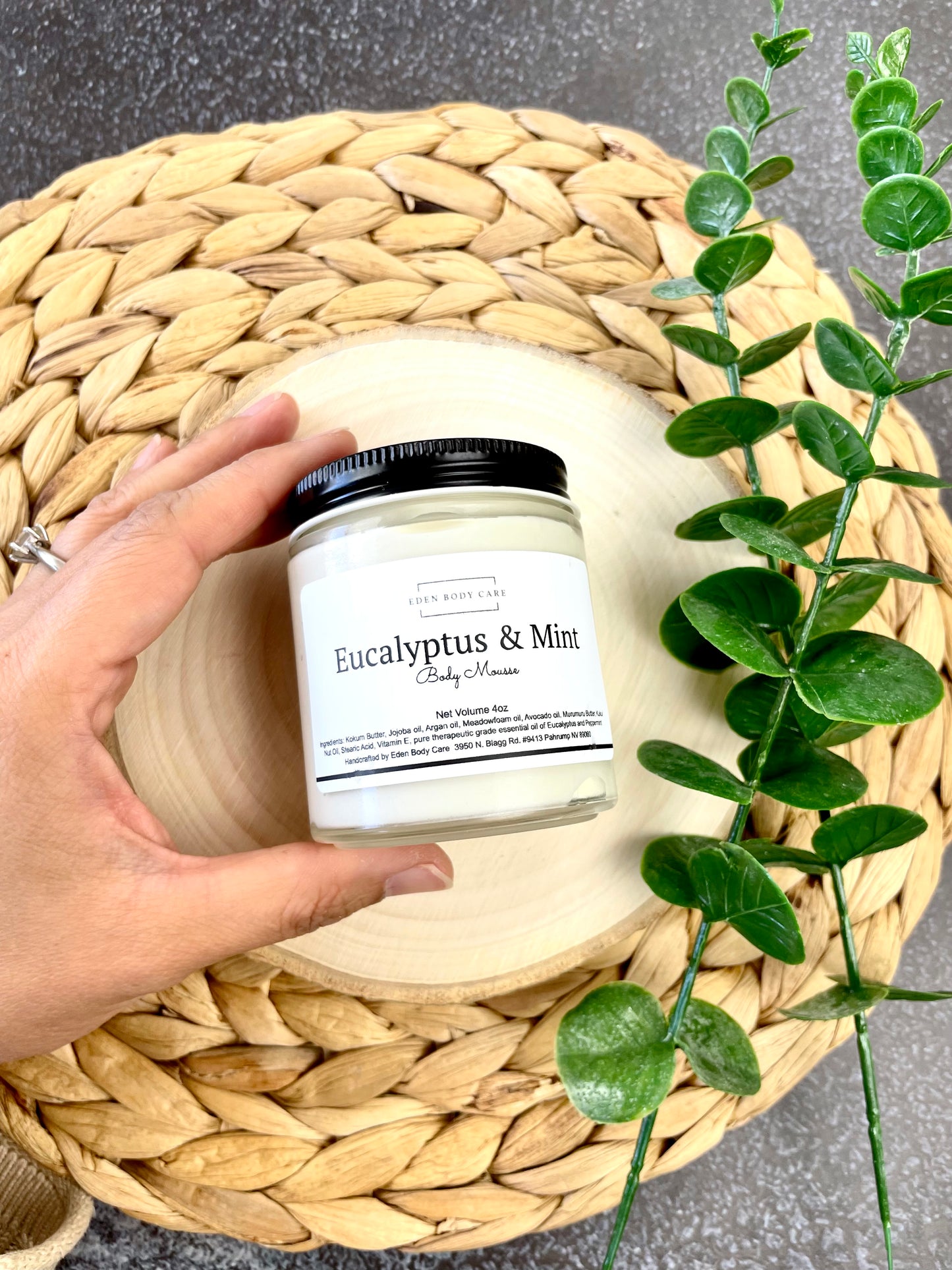 Eden Body Care Eucalyptus & Mint body mousse jar held in hand on a natural wood slice beside green leaves