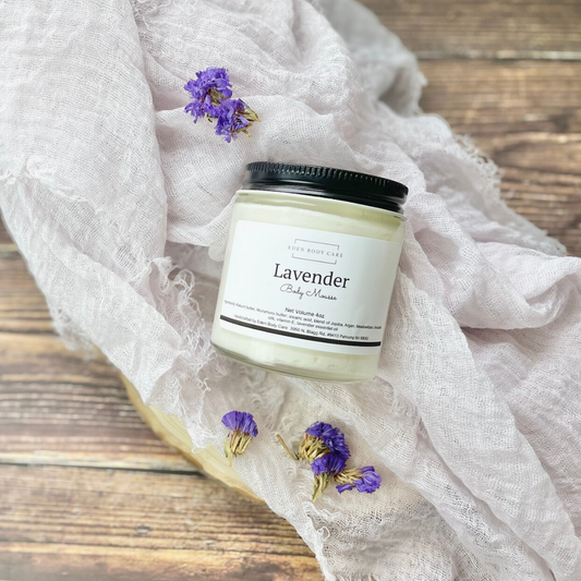 Eden Body Care Lavender body mousse in a glass jar displayed on soft white fabric with scattered purple flowers.