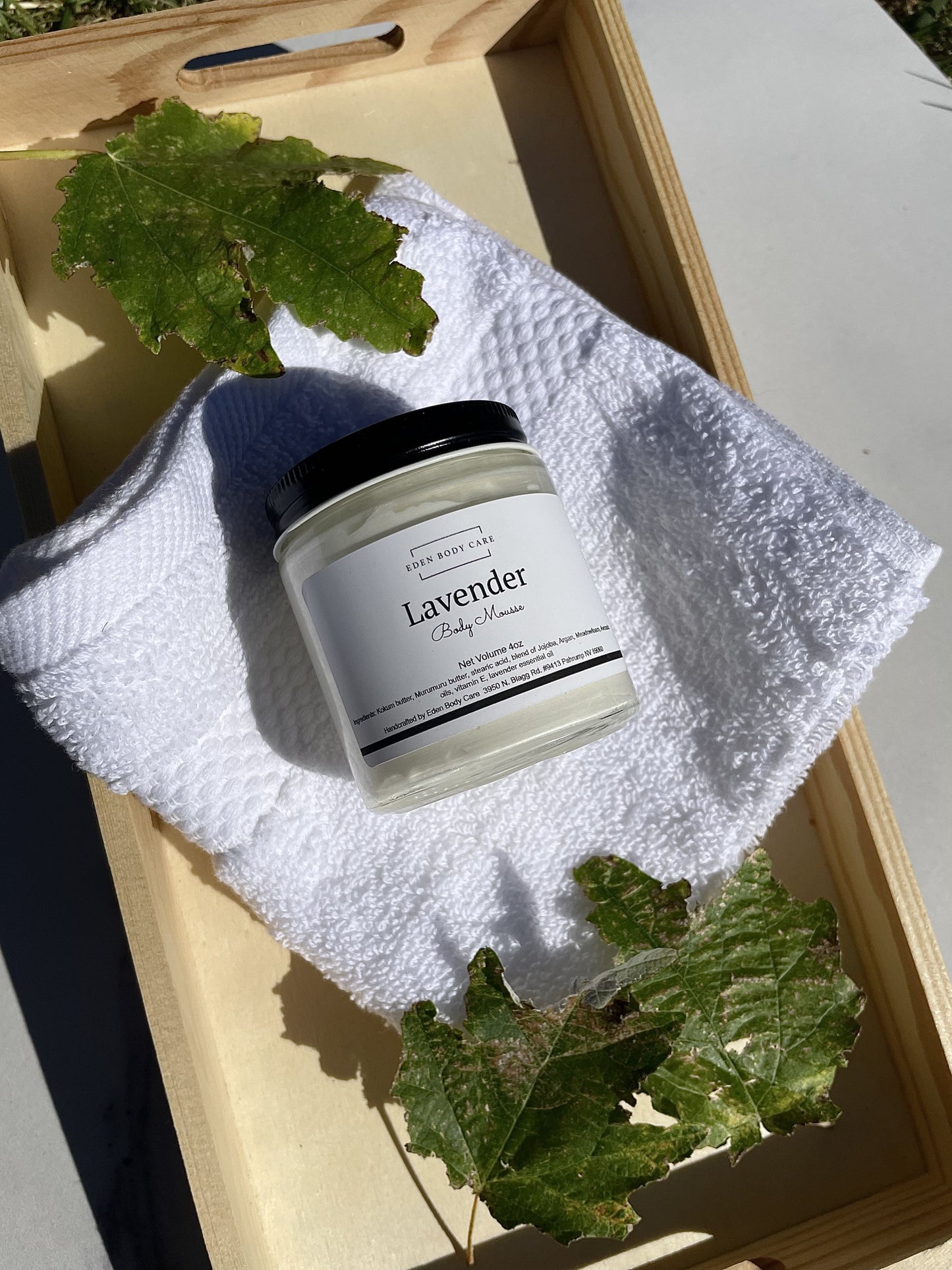 Eden Body Care Lavender body mousse jar styled on a white towel in natural sunlight with green leaves around it.