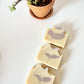 Three handmade goat milk soap bars with soft lavender botanical swirls displayed in a row beside a small potted plant.