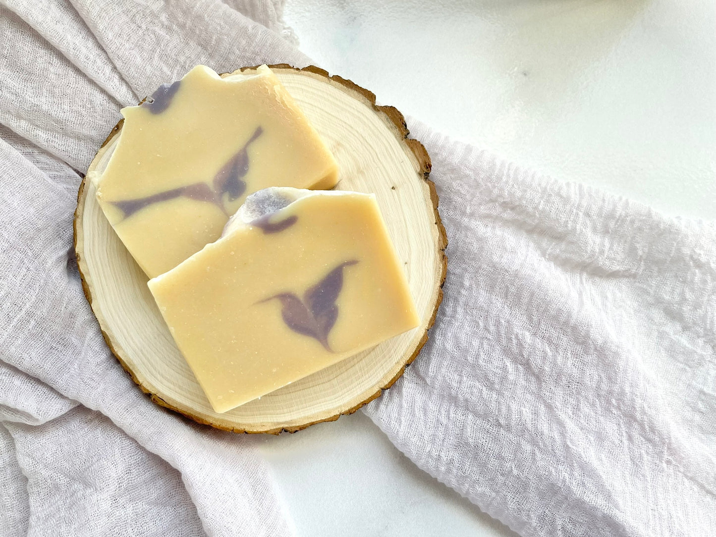 Two handmade goat milk soap bars with soft purple swirls displayed on a natural wood slice over neutral fabric.