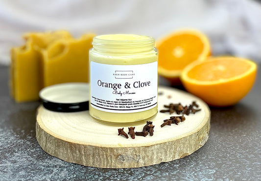 Eden Body Care Orange & Clove body butter displayed on a natural wood slice with fresh orange halves, whole cloves, and matching soap bars in the background.