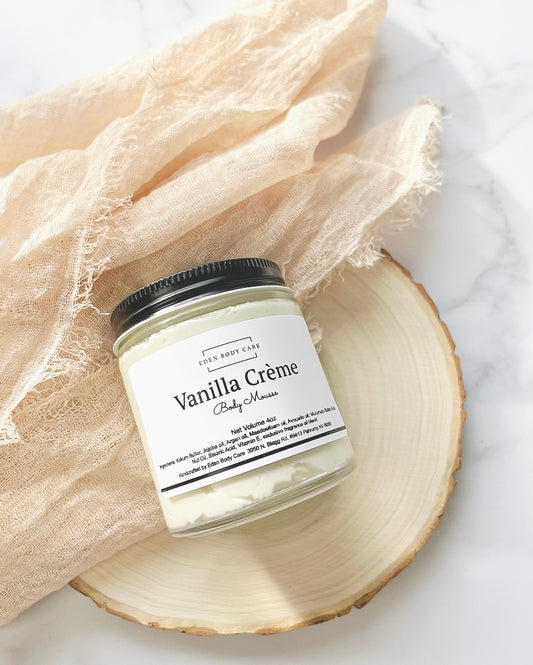 Eden Body Care Vanilla Crème body mousse jar displayed on a natural wood slice with soft neutral fabric in the background