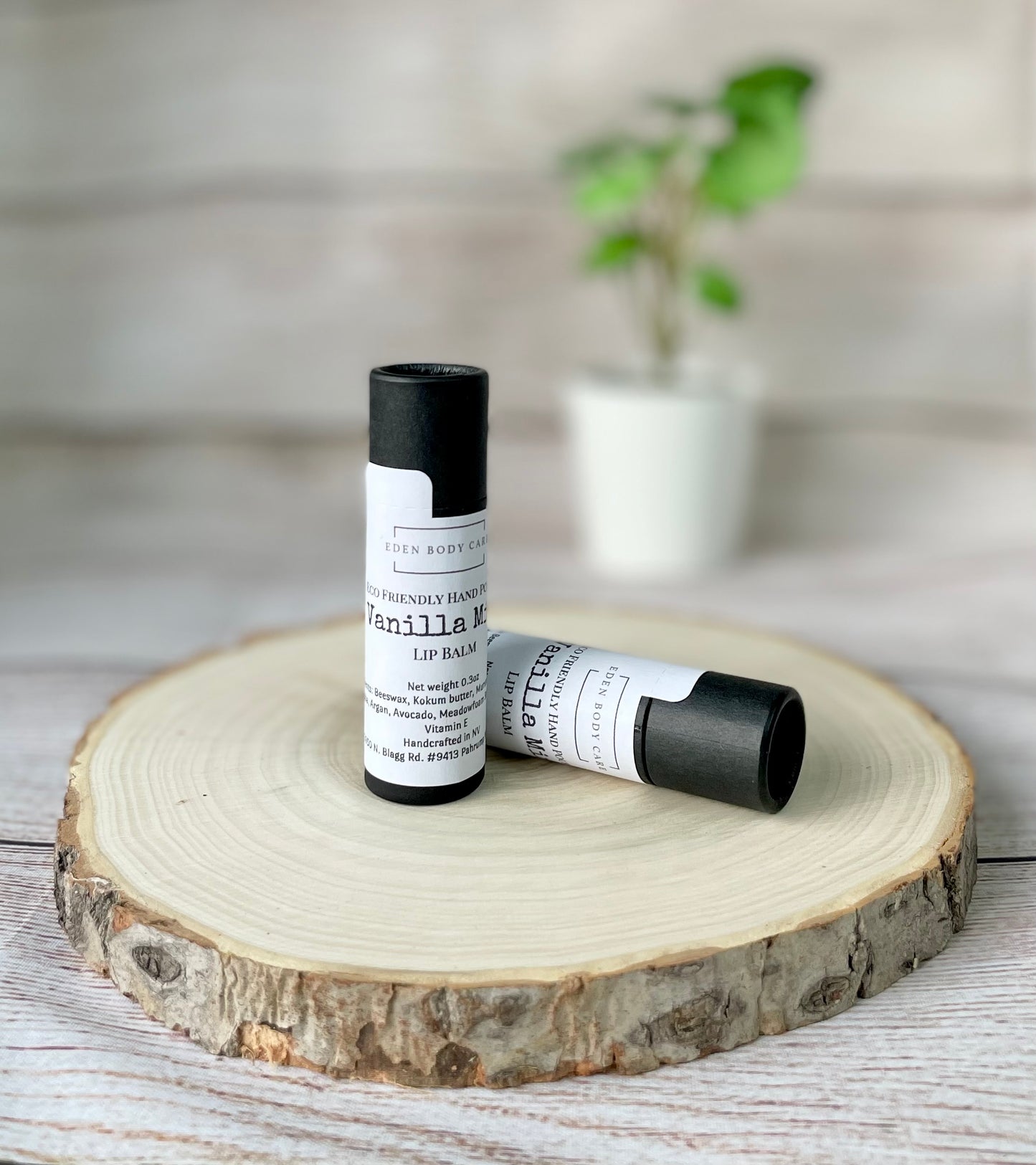 Two Eden Body Care Vanilla Mint lip balm tubes displayed on a natural wood slice with a softly blurred plant in the background.